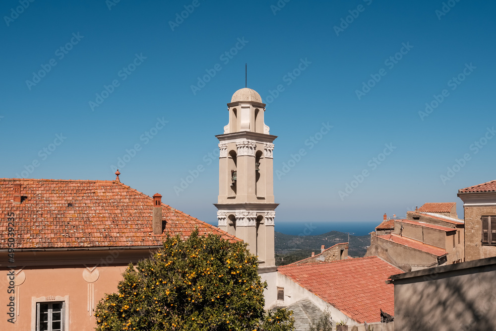The bell tower and terracotta roof tiles in the village of Costa in the Balagne region of Corsica with an orange tree in the foreground and Mediterranean sea in the distance