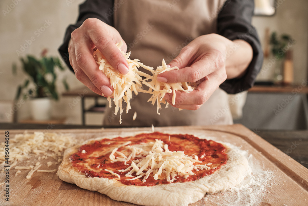 Close-up of unrecognizable woman standing at wooden board and sprinkling cheese on pizza dough with tomato sauce