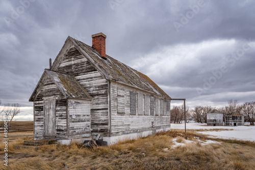 The exterior of an abandoned schoolhouse with two grain bins in the background on the prairies near Battrum, Saskatchewan © Nancy Anderson