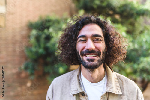 Positive Hispanic male with curly hair and beard smiling and looking at camera. While standing on blurred background of brick wall and lush tree on street