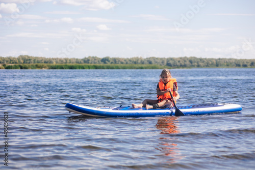 Female child sup boarding on her own on calm lake with oar in hands looking at water ripples in vest life jacket. Active holidays. Inculcation of love for sports from childhood.