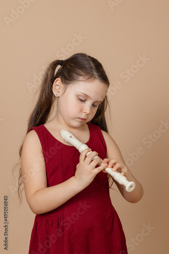 Girl in red dress learn melody by pinching holes in flute with bowed head, beige background. Tuition to play woodwind musical instrument. Flute and children is concept of music education development. photo