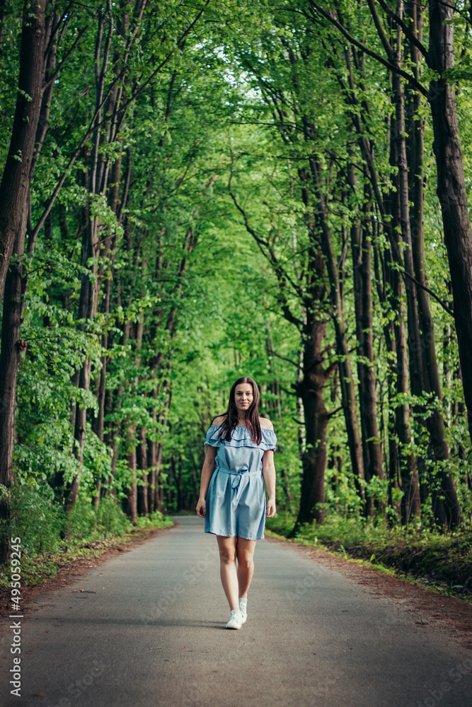 Fototapeta premium Young 20-24 year old slim brunette in a light blue summer dress dances on the road to the rhythm of the music. Candid portrait of a real ordinary person enjoying movement and dancing. Knowing oneself