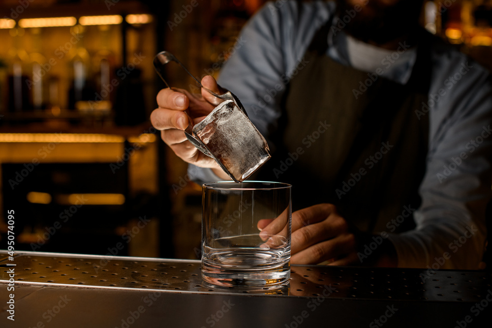 selective focus on big ice cube held by tongs in male hand over transparent glass