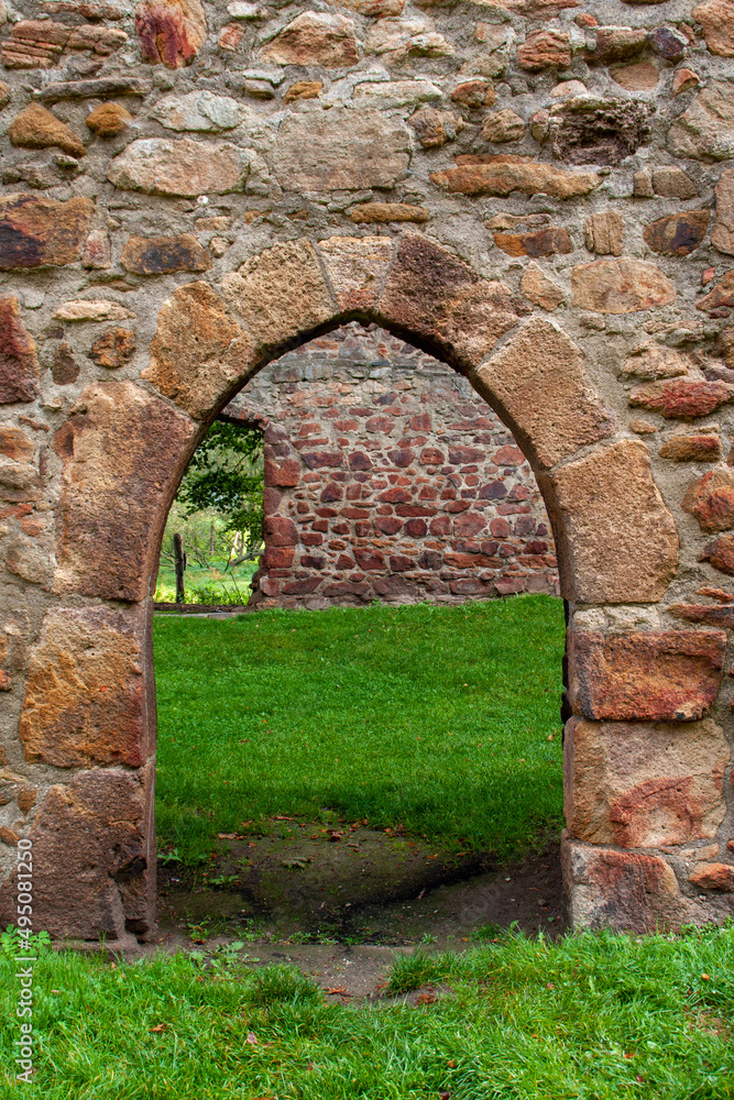 Fototapeta premium Passage through an old sandstone wall in the form of a pointed arch in a ruined monastery with deep green grass on the ground