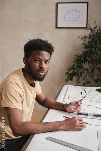 Portrait of serious frowning young African-American engineer with beard sitting at table with floor plans and holding eyeglasses