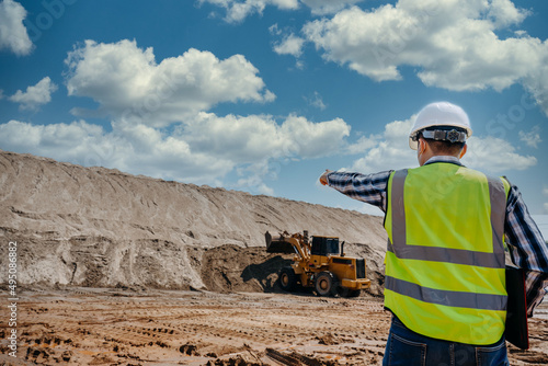 A young man civil engineer working at sand quarry inspects the operation of yellow excavators and sand dump trucks in the sandpit industry for use in construction at the construction site.