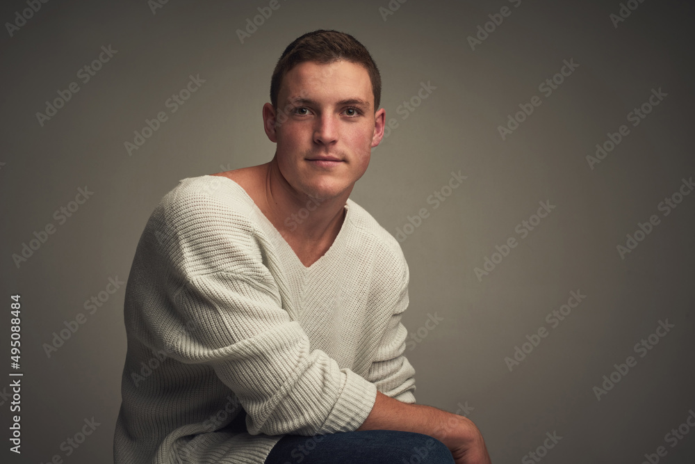 He can looked right through you. Studio portrait of a confident young man seated against a grey background while looking at the camer.