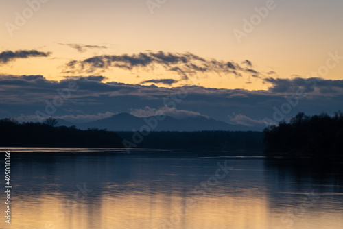 Landscape with Sava river and riparian zone forest silhouette, fog raises from distant mountain against dark clouds and orange glow in sky at dusk