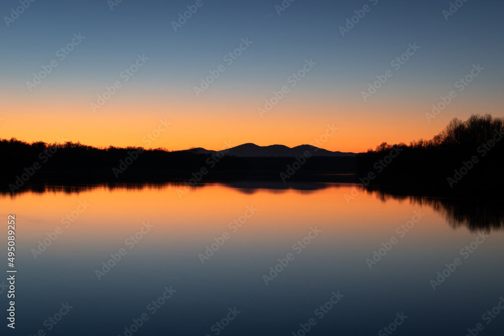 Landscape of mountain silhouette and forest with symmetric reflection in river water against clear blue sky with orange glow at horizon at twilight