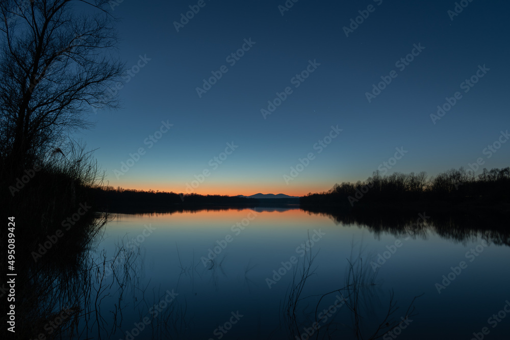 Landscape of mountain silhouette and forest at riverbank with reflection in river water against clear sky with first stars and orange glow at horizon at twilight