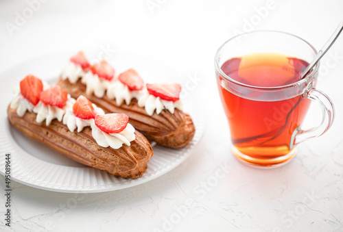Eclairs with cream and strawberries and a cup of tea on a white plate. 