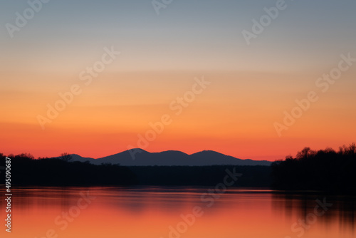 Landscape of Sava river, forest and distant mountain silhouettes, clear sky with vibrant red and orange glow at horizon during twilight