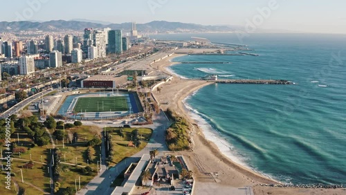 Extreme wide aerial shot of densely populated big modern city located on sea cost. Camera flying above residential areas, roadways, park and stadium to sandy seashore bathed in waves. Barcelona, Spain