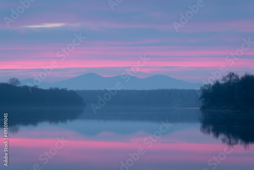 Fototapeta Naklejka Na Ścianę i Meble -  Peaceful atmospheric landscape with pastel colors, Sava river at twilight, forested banks lead to distant mountain silhouette in haze, calm nature and water reflection