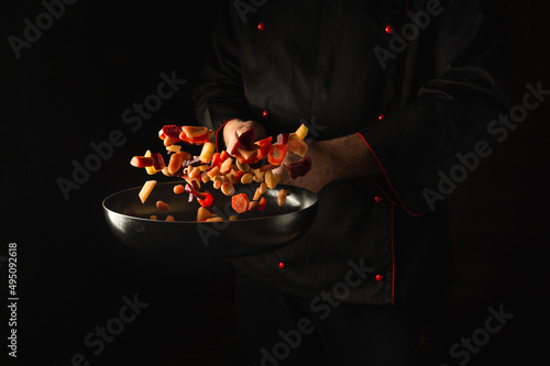 The chef prepares fresh vegetables in a frying pan. Grande cuisine and food recipe on dark background. Free advertising space