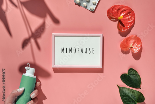 Menopause, text in pink frame. HRT Replacement hormone therapy concept. Pink background with exotic leaves, flowers, pills, estrogene gel in hand. Sunlight, long floral shadows. photo