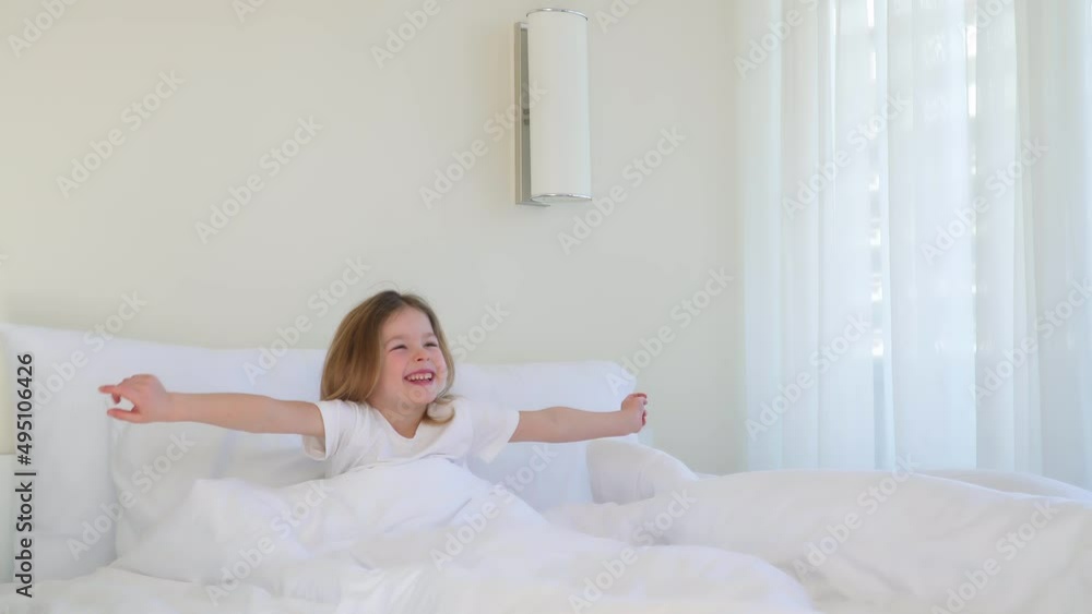 The child woke up. A little girl wakes up in a big white bed at home or in a hotel. Good morning happy person. Children's smile in the room. The concept of joyful childhood. Pull your hands. Stretch. The child woke up. A little girl wakes up in a big white bed at home or in a hotel. Good morning happy person. Children's smile in the room. The concept of joyful childhood. Pull your hands. Stretch.