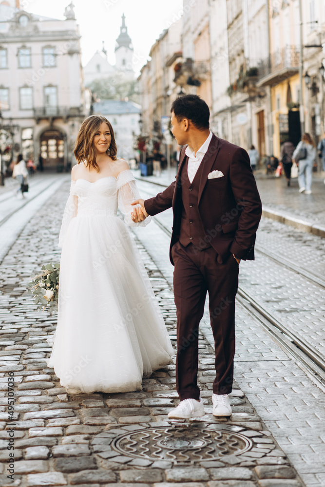 Naklejka premium An international wedding couple, a European bride and an Asian groom walk around the city together.