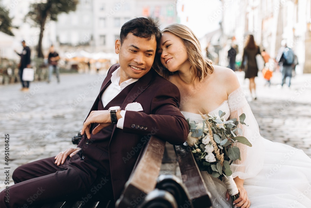 Fototapeta premium An international wedding couple, a European bride and an Asian groom walk around the city together.