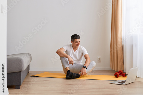 Horizontal shot of smiling Caucasian man wearing sportswear doing sport exercises at home, sitting on floor on yoga mat, looking at laptop screen, listening instruction online before workout.