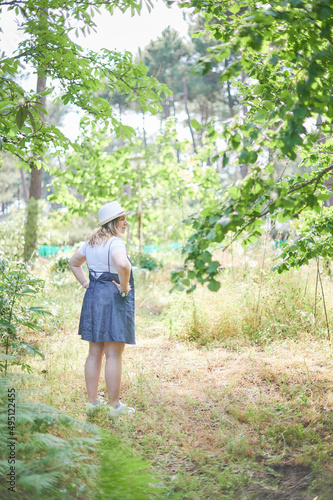 Young adult blonde curvy woman stands in the middle of the forest on a sunny day of summer. She is wearing a jeans dress, white shirt and a hat.
