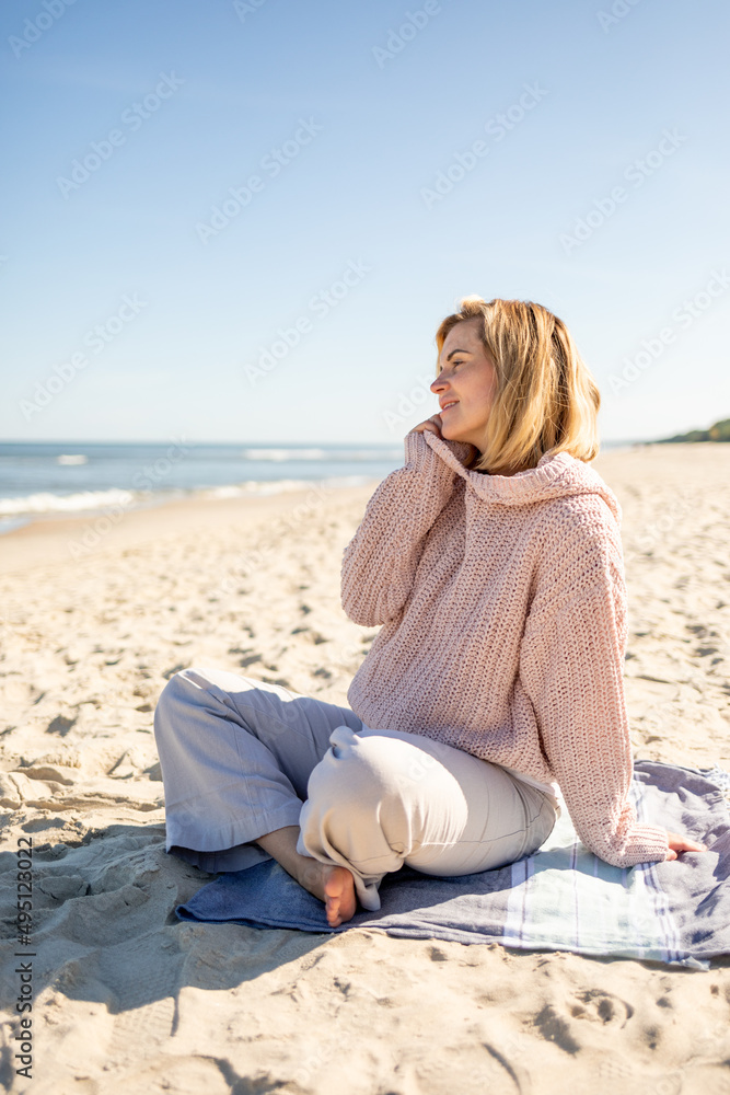 Charming young woman in knitted sweater sitting on sandy beach at sunny day
