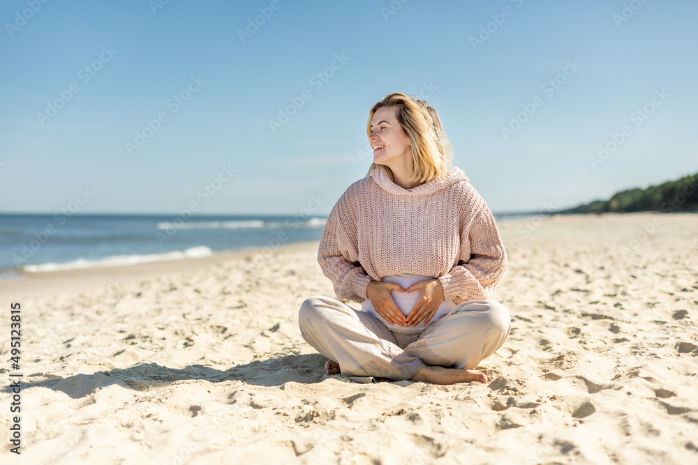 Pregnant woman in sweater sitting on seashore and relaxing