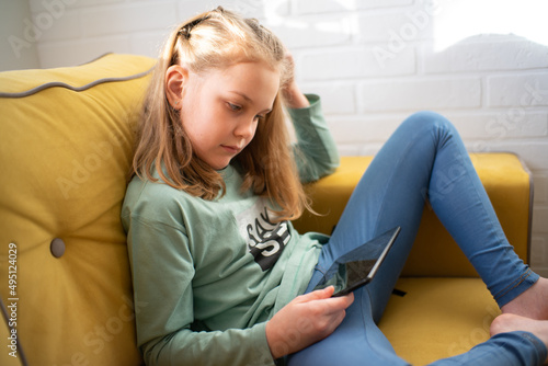 cute little girl is sitting on a sofa and enjoying playing an online game on a digital tablet computer, or listening to an online webinar, or reading a book.