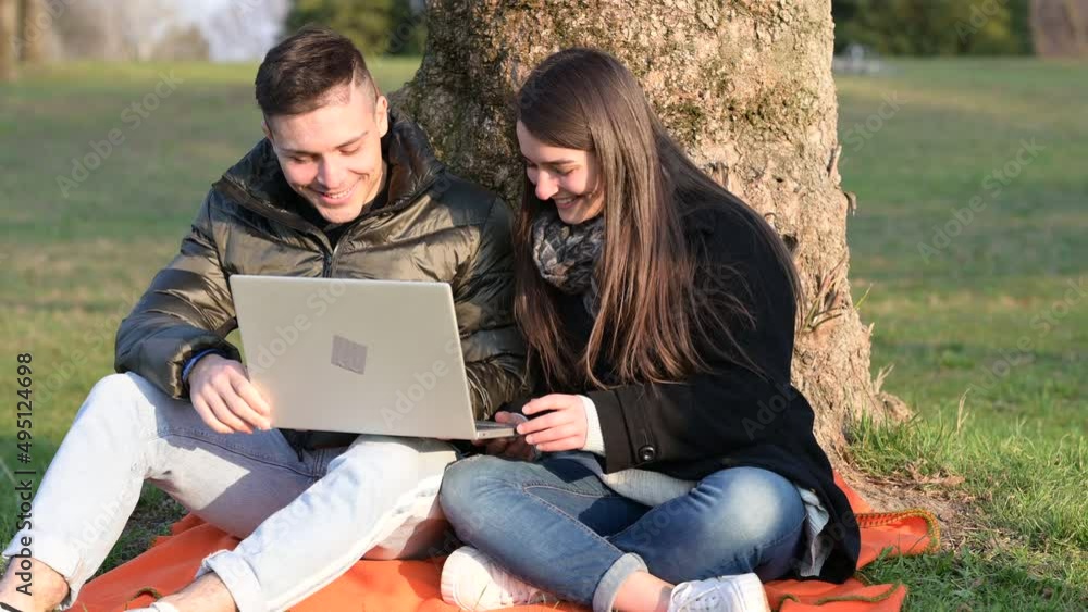 A beautiful couple in casual wear is having fun with the laptop outdoors. Sitting at the foot of a tree, you enjoy nature on a sunny spring day. Remote teamwork concept. A beautiful couple in casual wear is having fun with the laptop outdoors. Sitting at the foot of a tree, you enjoy nature on a sunny spring day. Remote teamwork concept.