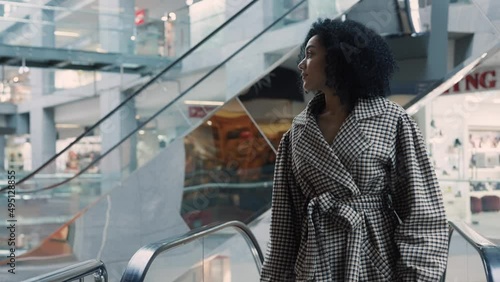 Stylish African American woman with colorful shopping bags in their hands climbs the escalator in the mall. Shopping, fashion and retail concept. Female shopper walks among shopwindows. Slow motion. photo