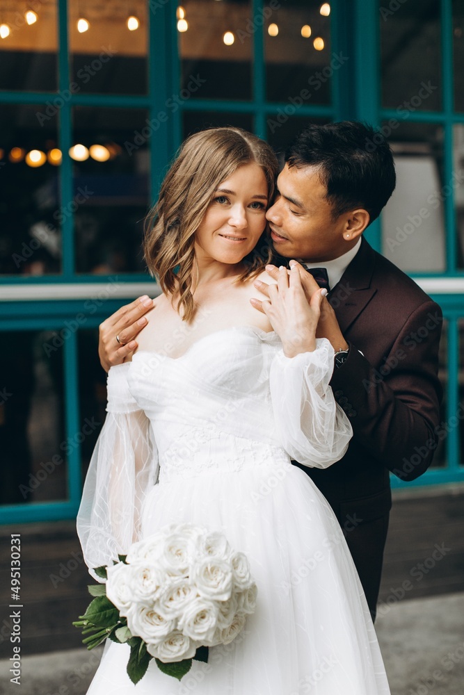 Naklejka premium Wedding day. Beautiful European bride and her Asian groom posing against the backdrop of cafes and large windows.