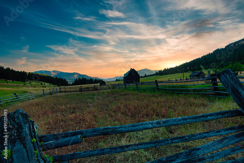 Early morning sunrays in orange and yellows with swirling clouds above a weathered barn, old fence and wide-open field at sunrise with rolling hills in the background – color graded