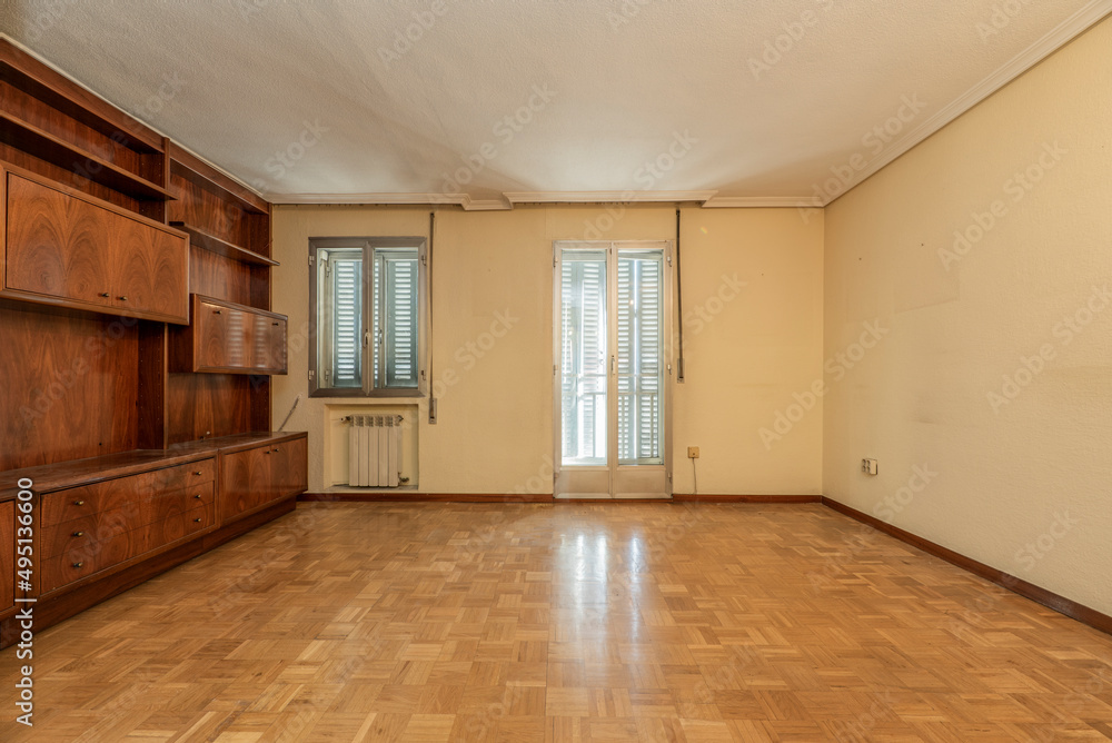 Wooden mural bookcase room, aluminum window and exit doors to covered terrace with radiator grill, cream-colored painted walls and plaster molding on the ceiling Wooden mural bookcase room, aluminum window and exit doors to covered terrace with radiator grill, cream-colored painted walls and plaster molding on the ceiling