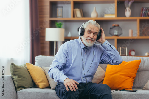 Senior gray-haired man sitting on sofa at home, relaxing and listening to music on headphones, using music app on phone