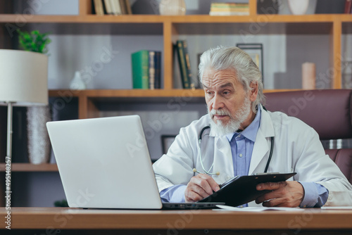Gray-haired experienced male doctor working on laptop remotely advising patients while sitting in office