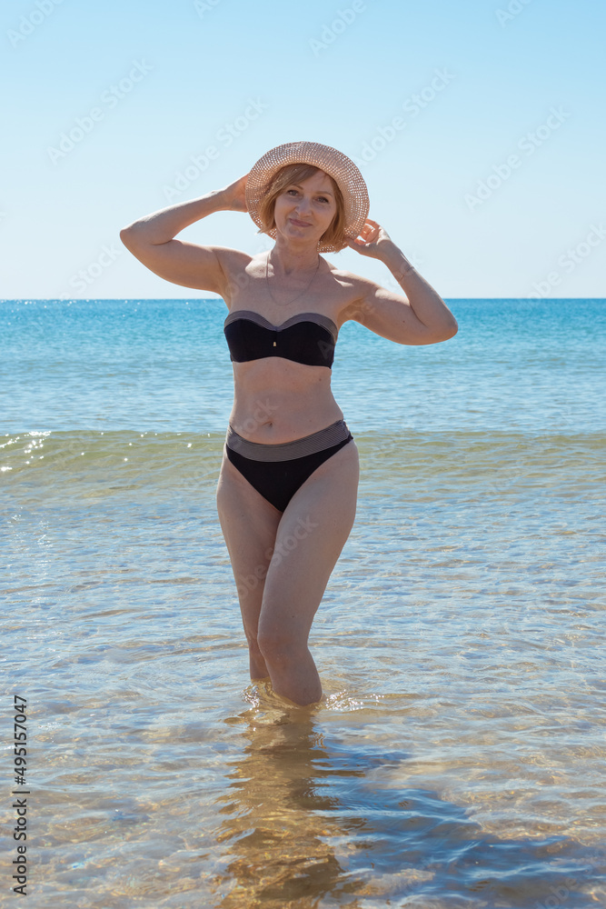 A mature slender woman in a swimsuit and a hat is standing in the sea water, smiling. Sea vacation and travel. A mature slender woman in a swimsuit and a hat is standing in the sea water, smiling. Sea vacation and travel.