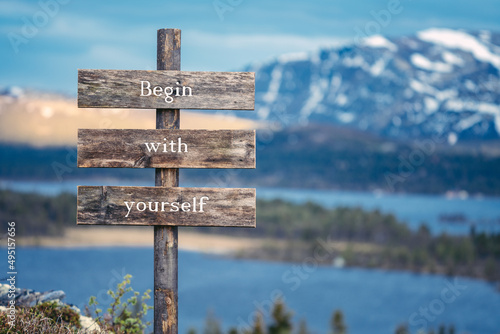 begin with yourself text quote written on wooden signpost outdoors in nature with lake and mountain scenery in the background. Moody feeling. photo