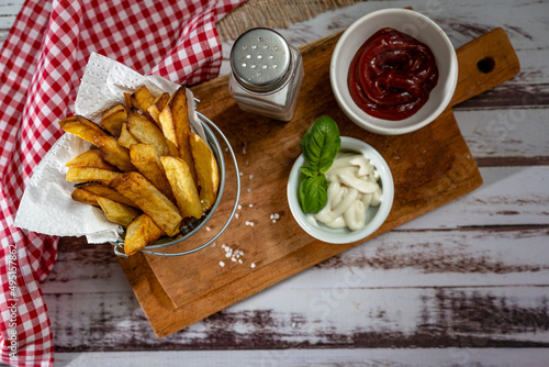 French fries in a metal pot with aioli and ketchup on a wooden board.
