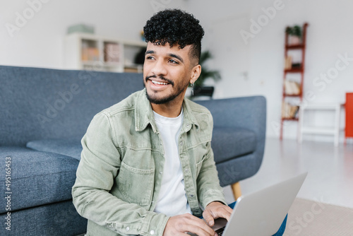 Casual young latino american man working on laptop sitting on floor at home - Millennial student working on computer in living room - Education and technology concept