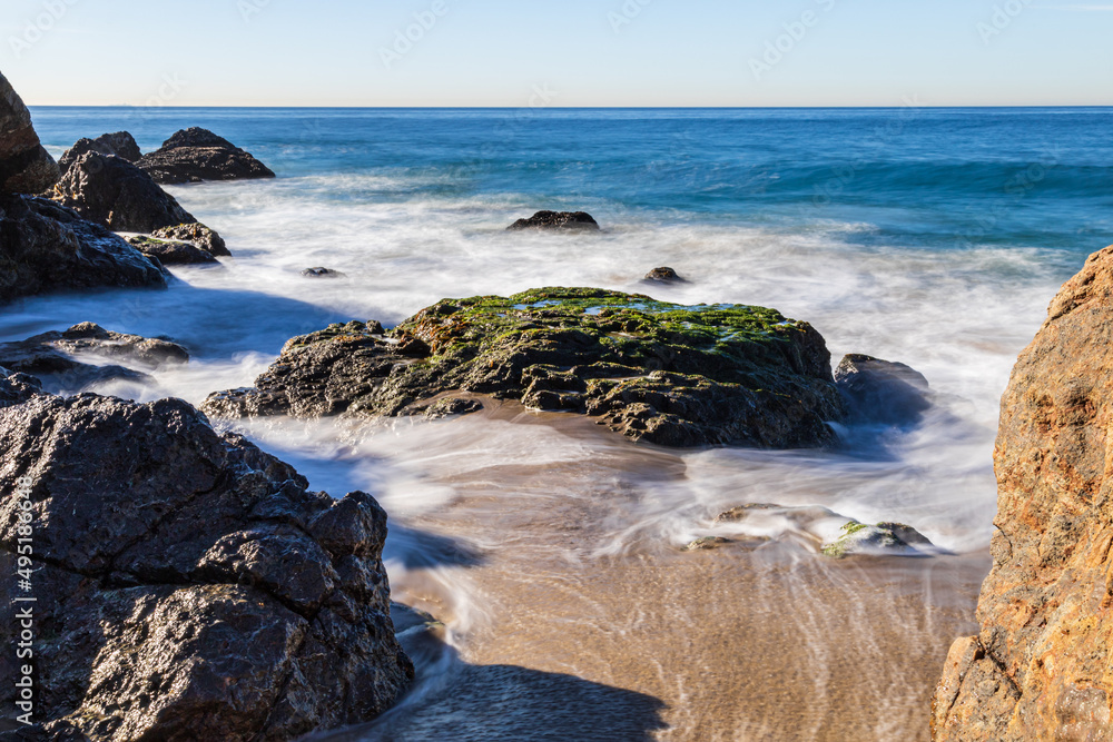 Rocky beach at Malibu, California. Waves receding from the sandy shore; blue Pacific Ocean in the background. 
