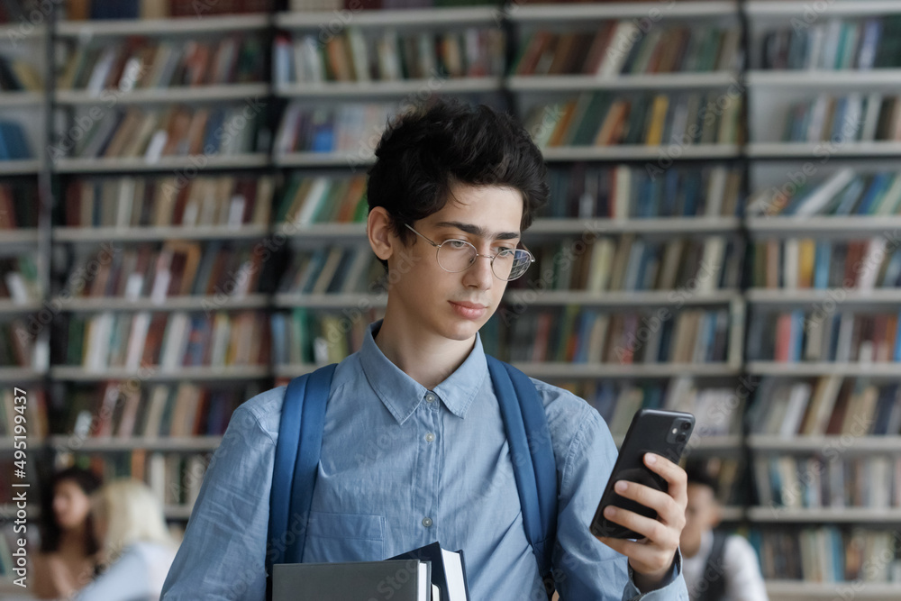 Student guy in glasses holds workbooks and cellphone check messages, chatting online standing in university library. Gen Z using modern tech and new mobile application, class timetable apps concept Student guy in glasses holds workbooks and cellphone check messages, chatting online standing in university library. Gen Z using modern tech and new mobile application, class timetable apps concept