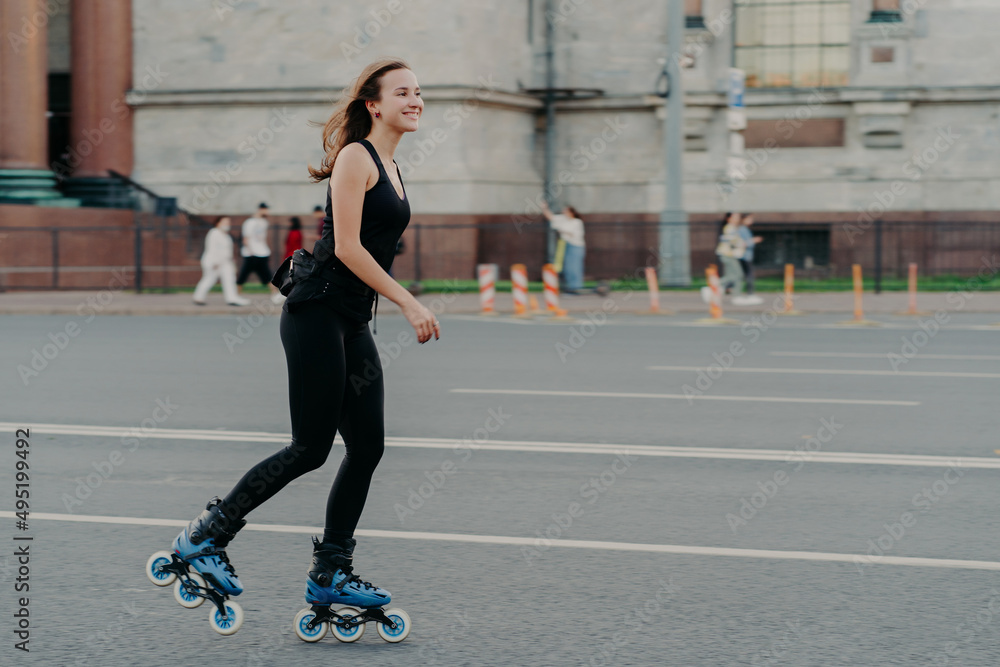 Athletic woman on roller skates moves actively poses on asphalt at street dressed in active wear has cheerful expression