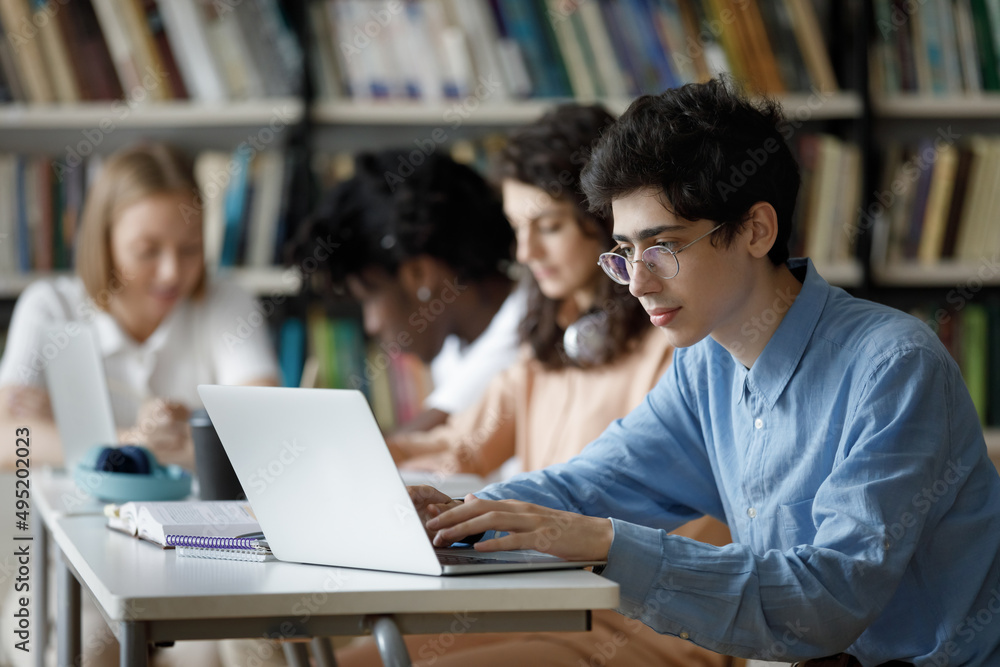 Student guy wear glasses studying in library or classroom, using laptop working on essay, prepare for college exams seated at table with diverse group mates. Education, professionals skills concept Student guy wear glasses studying in library or classroom, using laptop working on essay, prepare for college exams seated at table with diverse group mates. Education, professionals skills concept