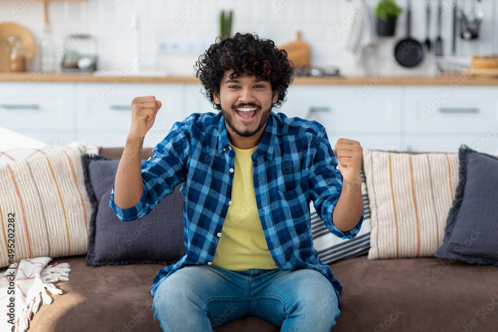 Portrait of happy Indian man overjoyed with good news. Smiling male with curly hair showing yes gesture, sitting on couch at home Portrait of happy Indian man overjoyed with good news. Smiling male with curly hair showing yes gesture, sitting on couch at home