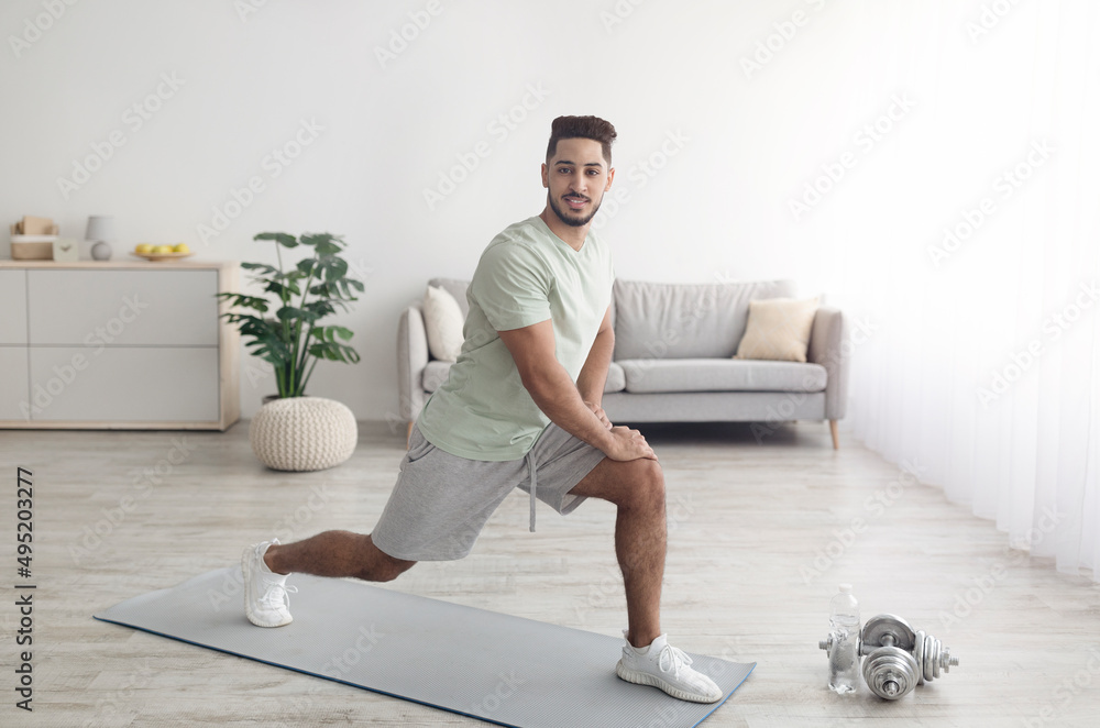 Fit young Arab guy making lunge, doing strength exercises, working out legs muscles at home, copy space