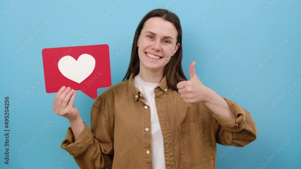 Cute smiling young woman holding heart icon, recommending to follow and share blog with interesting content, posing isolated over blue color background wall in studio. Like button of social media Cute smiling young woman holding heart icon, recommending to follow and share blog with interesting content, posing isolated over blue color background wall in studio. Like button of social media