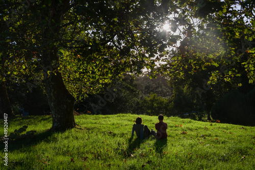 Couple sitting on the grass, sunbursts shining through the tree leaves, Auckland. © Janice
