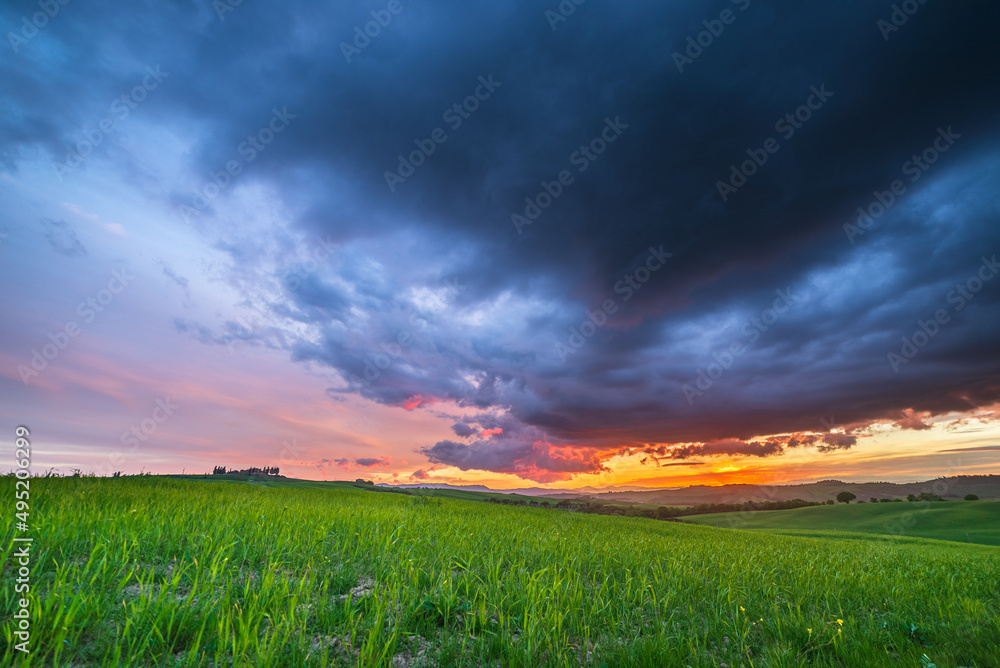 Unique green landscape in Tuscany, Italy. Dramatic sunset sky over cultivated hill range farm lands and cereal crop fields.