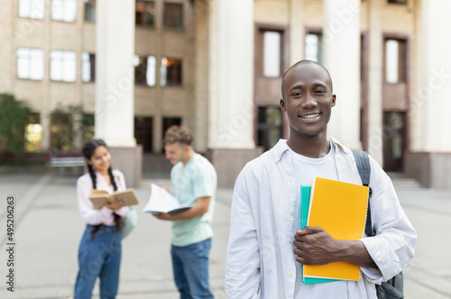 Male african american college student with backpack and workbooks outdoors, posing near university building